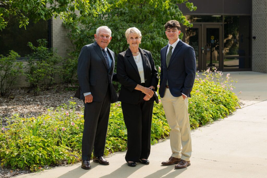 Three individuals in formal attire stand together outdoors on a paved path beside greenery and a building entrance.