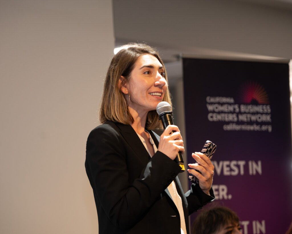 A person in a black blazer speaks into a microphone while holding a smartphone, standing before a banner for the California Women's Business Centers Network.