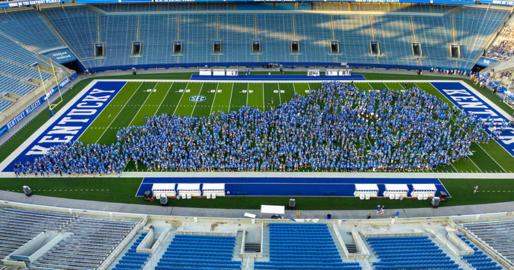 A large group of people in blue attire form a shape on a football field at a stadium with “KENTUCKY” written on the sidelines.